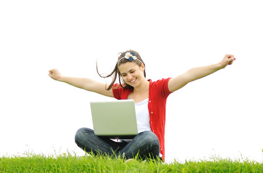 Kvinne sitter på en gressplen med laptop i fanget og strekker armene ut fra kroppen.
